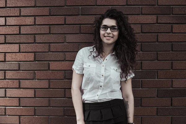 Mutlu genç kadın kırmızı tuğla duvar arka plan kameraya poz. Uzun siyah kıvırcık saçlı kız beyaz bluz giyen gözlük ve saat giyinmiş. Kameraya bakarak ve gülümseyen kadın