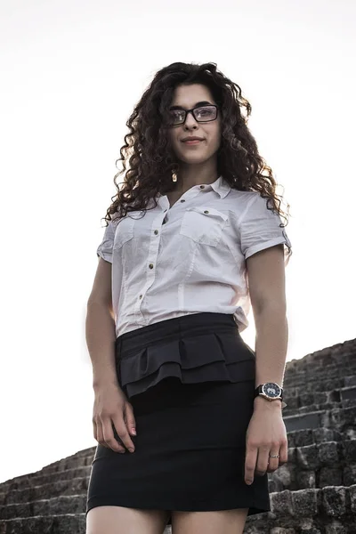 Woman in white short blouse and in glasses standing on the stairs and looking at camera. Mixed race girl with pretty face. Outdoor. Portrait. Full-length