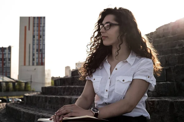 Genç ve oldukça karışık ırk kadın beyaz bluz, siyah etek ve merdivenlerde oturan ve uzağa bakarak gözlük. Belgili tanımlık defter tutan eller kız.
