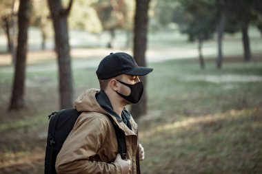 Tıbbi maskeli adam parkta yürüyor. Sırt çantalı, ceketli ve temiz hava alan bir adam. 