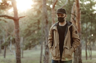 Siyah maskeli ve şapkalı bir adam ağaçların arkasında kameraya poz veriyor. Adam kameraya bakıyor. Sağlık hizmetleri. Epidemy