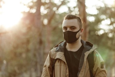Tıbbi maskeli portre adamı yakından çek. Genç adam ağaçların arkasında duruyor ve kameraya bakıyor. Açık havada. Sağlık ve Bakım. Virüs