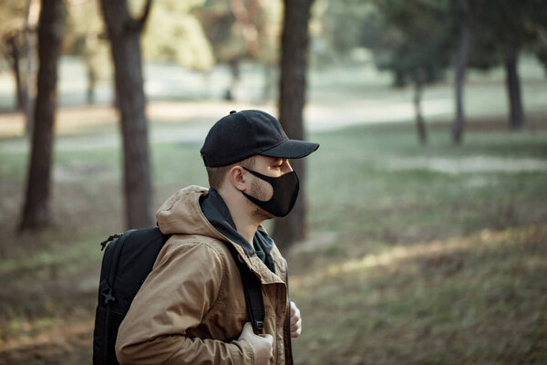 Человек в медицинской маске проходит через парк. Человек в рюкзаке в пиджаке и колпаке дышит свежим воздухом
 
