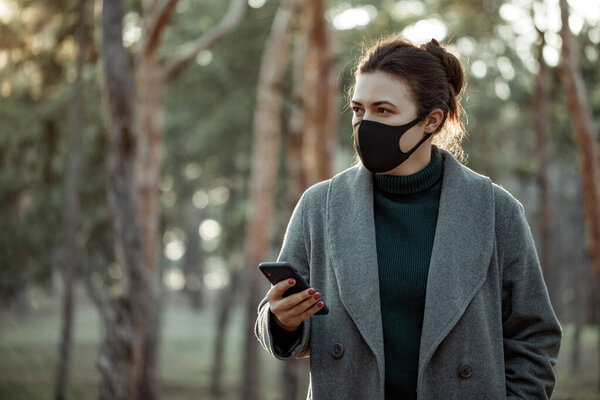 Молодая женщина в защитной маске со смартфоном. Девушка, стоящая на фоне деревьев и смотрящая в сторону
