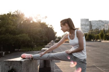 Beyaz kolsuz tişörtlü sportif Avrupalı kız bacaklarını esnetip ısınıyor. Dışarıda egzersiz yapan genç bir kadın.