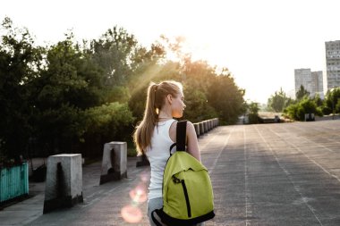 Beyaz kolsuz tişörtlü, sırtında sırt çantasıyla yolda yürüyen genç bir Avrupalı kızın arka görüntüsü.