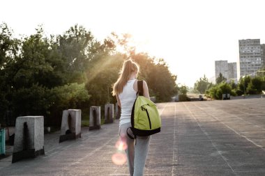 Beyaz kolsuz tişörtlü, sırtında sırt çantasıyla yolda yürüyen genç bir Avrupalı kızın arka görüntüsü.