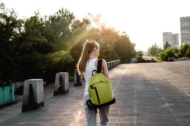 Beyaz kolsuz tişörtlü, sırtında sırt çantasıyla yolda yürüyen genç bir Avrupalı kızın arka görüntüsü.