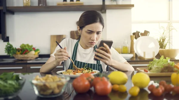 Modern Mutfakta Otururken Salata Yiyen Asyalı Kadınlar. Ve ciddi bir ifadeyle haberlere bakıyorlar. Evde Coronavirus salgını sırasında gözaltına alınırken (Covid-19).