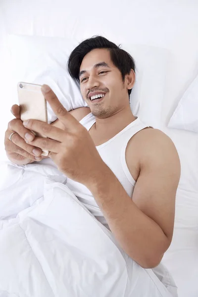 Hombre asiático acostado en la cama con teléfono inteligente —  Fotos de Stock