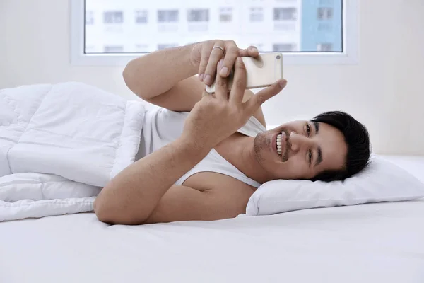 Азиатский мужчина лежит в постели со смартфоном — стоковое фото