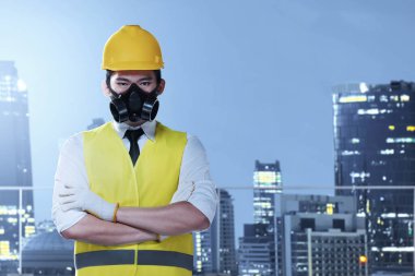 Korumalı maskeli ve sarı kasklı Asyalı işçi şehrin arka planında duruyor. İşçi Bayramı konsepti