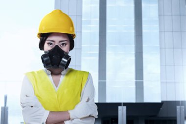 Korumalı maskeli ve sarı kasklı Asyalı bir kadın bina arka planında duruyor. İşçi Bayramı konsepti