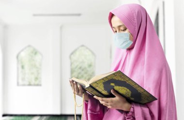 Duvak takmış, grip maskesi takmış elinde dua boncuklarıyla Kur 'an okuyan Asyalı Müslüman bir kadın. Grip salgını Coronavirüs 'ü önler.