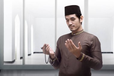 Asyalı Müslüman bir adam caminin içinde elinde dua boncuklarıyla dua ediyor.