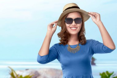 Şapkalı ve güneş gözlüklü Asyalı bir kadın mavi gökyüzü arka planıyla plaja gidiyor.