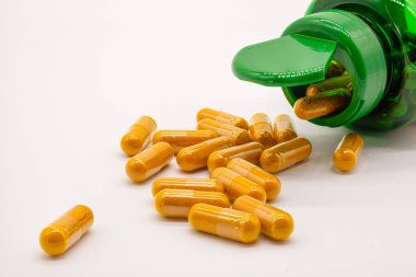Beyaz bir yüzey üzerinde zerdeçal tozu içeren plastik kapsüller ve yeşil plastik bir şişeden dökülen beyaz arka plan.