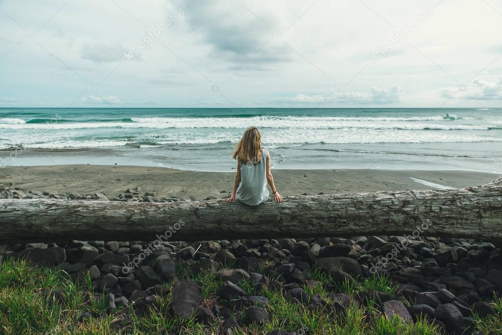 One girl sits on a large wood bench looking at the ocean and cloudy sky no face