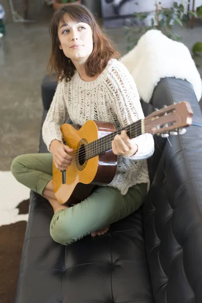 Hermosa joven sentada en el sofá tocando la guitarra — Foto de Stock