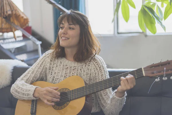 Belle jeune femme assise sur un canapé jouant de la guitare — Photo