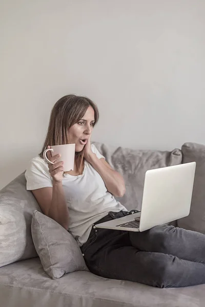 Schöne Frau überrascht beim Anblick eines Computerbildschirms, während sie eine Tasse Kaffee hält — Stockfoto