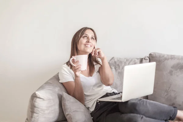 Belle femme adulte se détendre sur le canapé tenant une tasse de café tout en utilisant un ordinateur portable — Photo