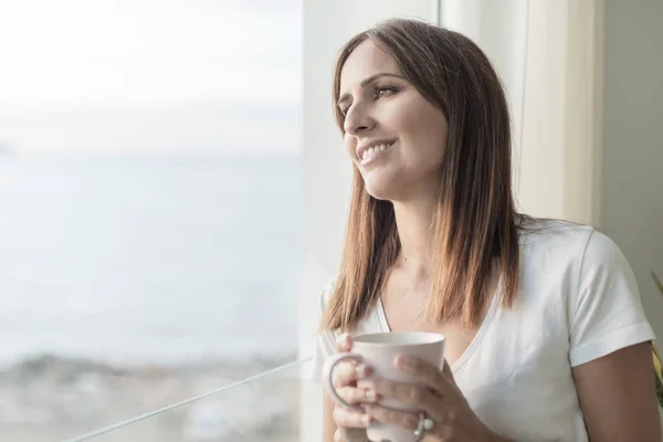 Mujer adulta de pie mientras mira por la ventana degustación de café — Foto de Stock