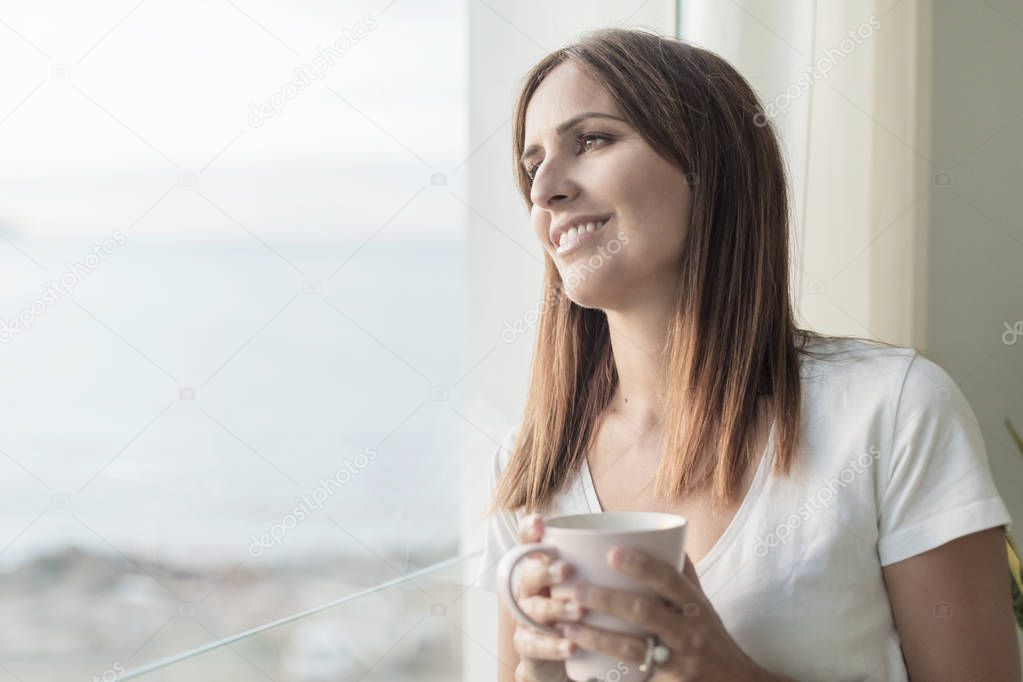 Adult woman standing while looking through window tasting coffee