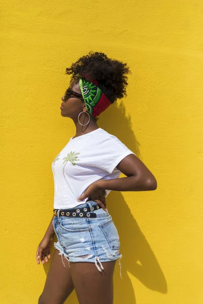 Vista lateral de uma mulher afro com óculos de sol e lenço de cabeça apoiados na parede amarela enquanto olha para o lado — Fotografia de Stock