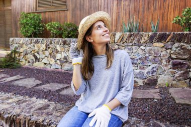 Güzel, genç bahçıvan kadın, şapkası ve eldivenleri ile avluda oturmuş gülümsüyor.