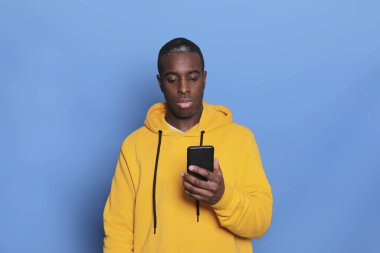 Sarı kazaklı neşeli Afro-Amerikan adam cep telefonu uygulaması kullanıyor. Mavi arka planda cep telefonu tutan mutlu hippi adam.