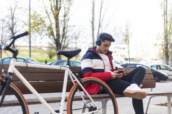 Fiatal férfi ül egy padon az utcán, miközben használja a mobiltelefon és zenét hallgat a fejhallgató — Stock Fotó