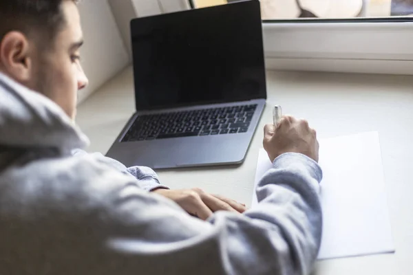 Achteraanzicht van een jongeman die thuis op papier schrijft naast een laptop — Stockfoto