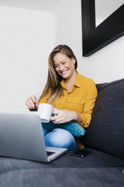 Oldukça uzun saçlı bir kadın portatif bilgisayarla koltukta oturuyor. Evden uzaktan bir fincan çayla çalışıyor.