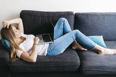 Kanepede oturan oldukça uzun saçlı bir kadın taşınabilir bilgisayarla evden uzaktan çalışıyor..