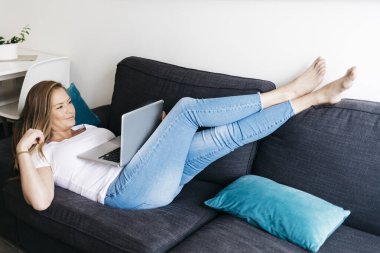 Kanepede oturan oldukça uzun saçlı bir kadın taşınabilir bilgisayarla evden uzaktan çalışıyor..