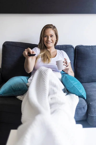 一个快乐的年轻女人坐在家里的沙发上 用遥控器 — 图库照片