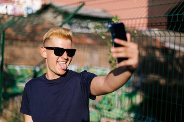 Güneş gözlüklü genç erkek dışarıda selfie çekiyor.