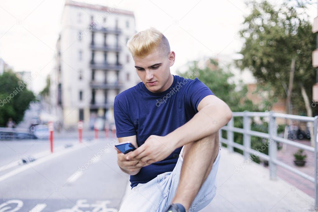 Young teenage boy with blonde bangs sitting on the street while using a mobile phone