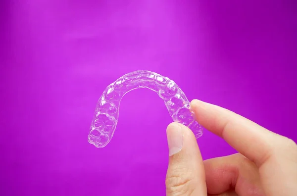 手握透明的塑料义齿 在紫色背景下隔离 是供牙齿矫正医生在牙科诊所或医院接受牙齿矫正手术的设备 — 图库照片