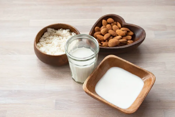 Almond milk in a glass, almonds, milk and squeezed nut in bamboo bowl on wooden background close up view. Selective soft focus. Text copy space. Homemade almond milk concept