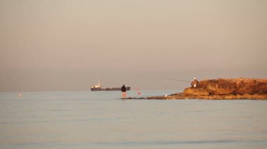 Bir balıkçı deniz şafak, şafak gökyüzü, kayaya üzerinde bir balıkçı siluetleri plaj balıkçılık çubuklar ile balıkçıların balık tutuyor. Şafakta deniz kıyısında balık adam. Denizde gemi.