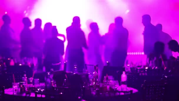 A youth party in a restaurant or a nightclub, banquet tables with alcohol and food against the background of silhouettes of dancing people, stage light and purple fill — Stock Video