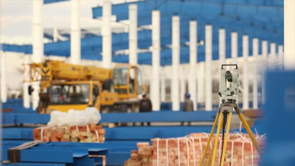 Teodólito óptico, Teodólito de construtores, teodólito de construção no fundo da construção de um grande edifício industrial ou armazém, trabalhos de construção — Vídeo de Stock