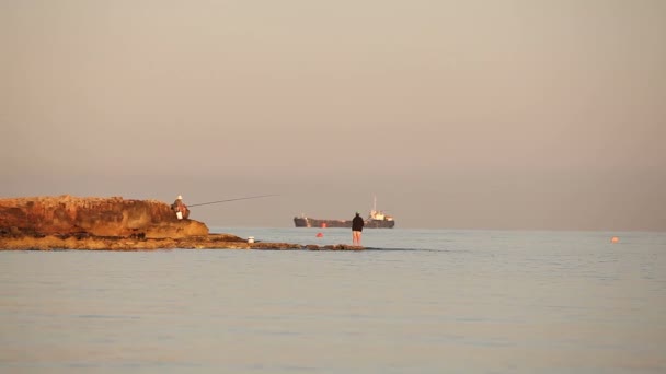 漁師は、ビーチで釣り竿と漁師のシルエット夜明けの海、夜明けの空を岩の上の漁師の釣りです。海岸の夜明けに釣り人。海を船します。. — ストック動画