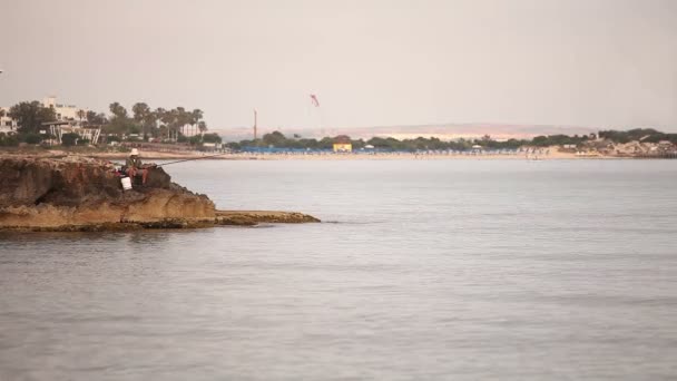 Un pescador está pescando en el mar al amanecer, un pescador en la roca contra el cielo del amanecer, siluetas de pescadores con cañas de pescar en la playa. Hombre pescando al amanecer en la orilla del mar. Nave en el mar . — Vídeos de Stock