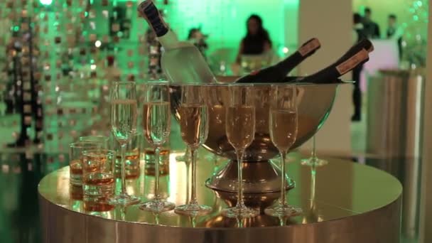 Copos de champanhe e vinho na mesa de buffet, uma garrafa de champanhe ou vinho em um balde em um fundo de pessoas dançando, silhuetas borradas de pessoas — Vídeo de Stock