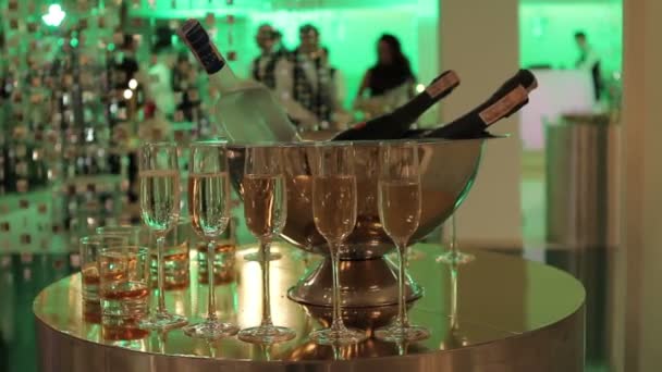Copos de champanhe e vinho na mesa de buffet, uma garrafa de champanhe ou vinho em um balde em um fundo de pessoas dançando, silhuetas borradas de pessoas — Vídeo de Stock