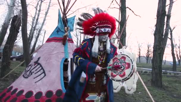 Північноамериканські індіанці в повному параді, людина в костюмі індійських танців з бубном на вулиці на очах Вігвам — стокове відео