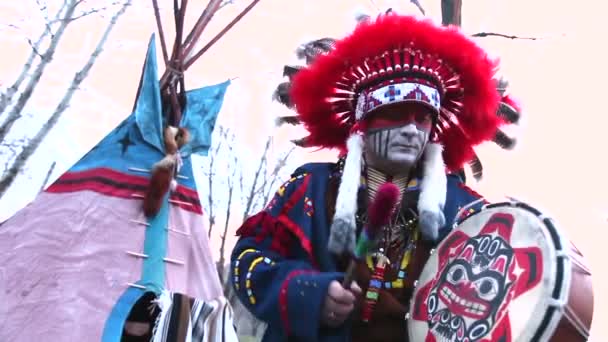 Indios norteamericanos vestidos de gala, un hombre con traje indio bailando con una pandereta en la calle frente a Wigwam — Vídeos de Stock